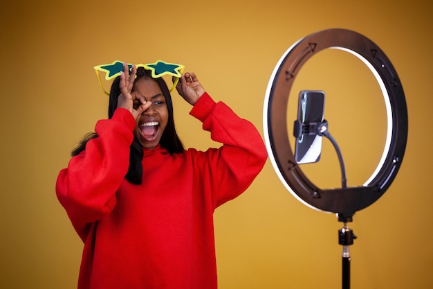 Laughing American blogger girl in a red hoodie and large star glasses with a lighted ring lamp