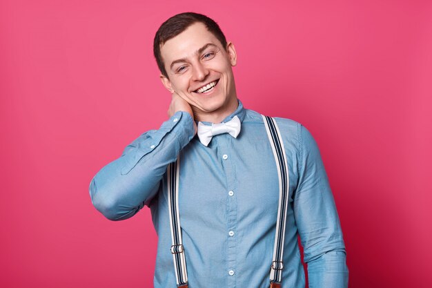 Laughing attractive boy putting one hand on neck, tilting his head on right side, wearing blue shirt with white buttons