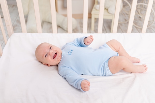 Смеющийся ребенок лежит в кроватке, милый маленький мальчик шести месяцев лежит в детской на кровати и смеется
