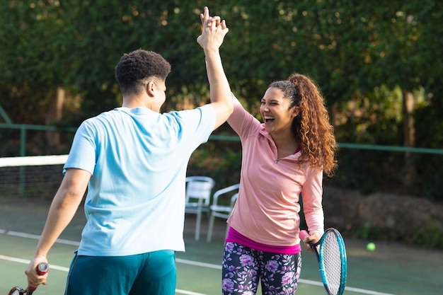 Laughing biracial couple playing tennis shaking hands on sunny outdoor tennis court. Inclusivity, sport, healthy hobbies, competition, fitness and leisure time concept.