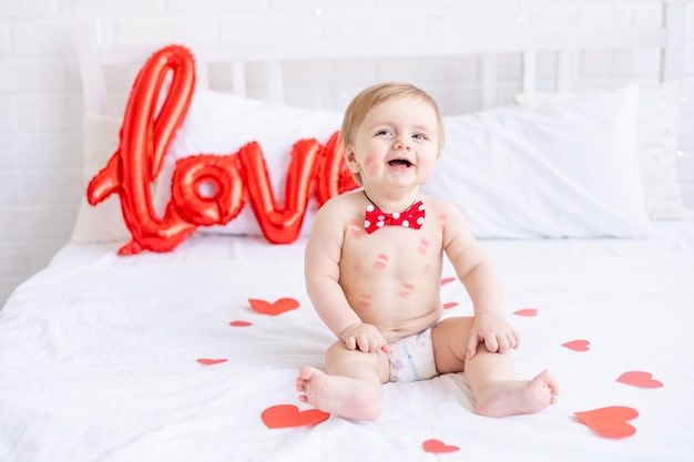 A laughing blonde baby boy is sitting on the bed in diapers with kisses from red lipstick at home and smiling among hearts and a large balloon with the inscription love the concept of Valentines Day