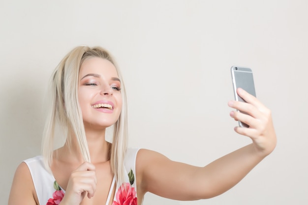 灰色の背景に対して携帯電話でselfieを作る金髪の女性を笑う