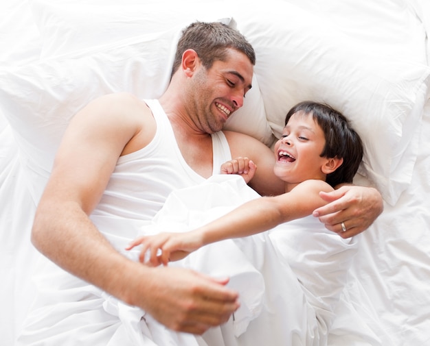 Смеющийся мальчик, играющий со своим отцом, лежащим на кровати у себя дома