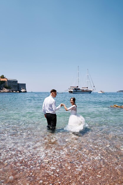 Foto la sposa e lo sposo ridono e entrano nelle profondità del mare tenendosi per mano e guardandosi intorno.