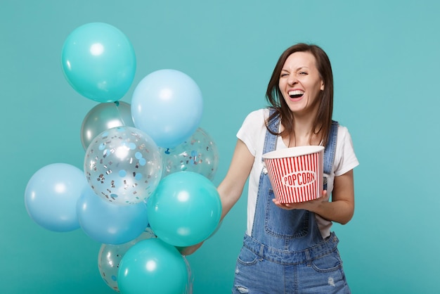 Laughing cheerful young woman in denim clothes holding bucket of popcorn, celebrating with colorful air balloons isolated on blue turquoise background. Birthday holiday party, people emotions concept.
