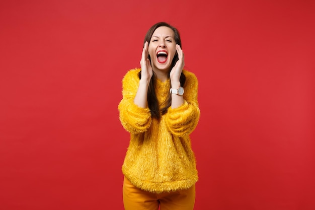 Laughing crazy young woman in yellow fur sweater screaming with hand gesture near mouth isolated on bright red wall background in studio. People sincere emotions lifestyle concept. Mock up copy space.