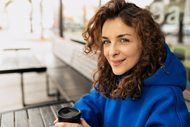 Laughing curly darkhaired girl looking at camera while sitting on old street in morning Pretty young lady with blue eyes posing with coffee waiting friend on avenue