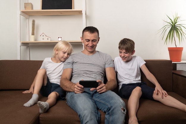 Laughing dad and two sons are sitting on couch and watching a video on phone.