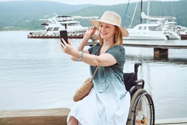 Laughing disabled woman in a wheelchair makes a selfie on the background of sea yachts