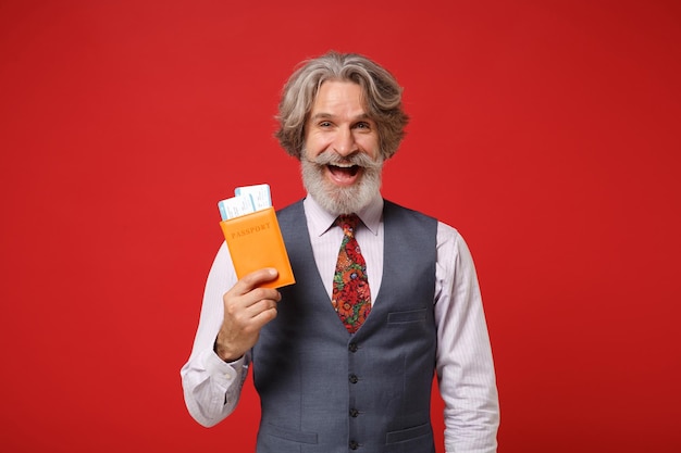 Laughing elderly gray-haired mustache bearded man in classic shirt vest colorful tie isolated on red background. People lifestyle concept. Mock up copy space. Holding passport boarding pass ticket.
