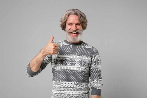 Laughing elderly gray-haired mustache bearded man in knitted sweater posing isolated on grey wall background in studio. People sincere emotions lifestyle concept. Mock up copy space. Showing thumb up.