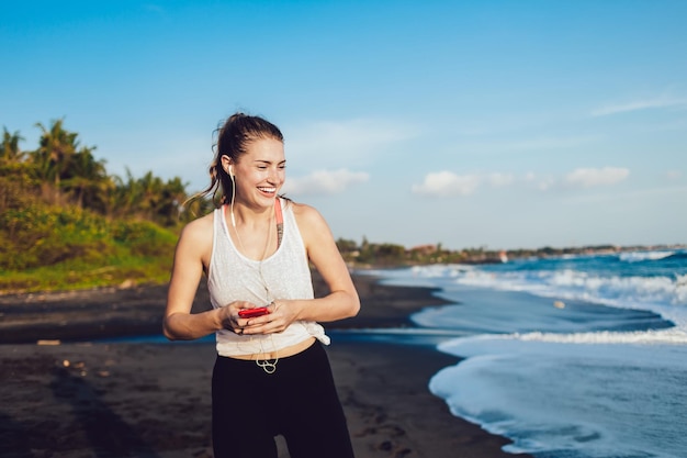 Laughing female jogger in earphones messaging on mobile and listening to music on beach