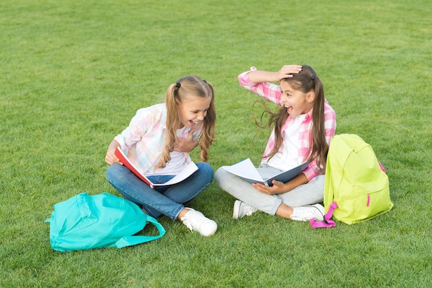 冗談から笑う。緑の芝生で楽しんでください。バックパックを持つ2人の小さな子供。小さな女の子は学校の休憩で遊んで勉強します。幸せな子供の日。姉妹関係と家族の概念。知識の日が来ました。