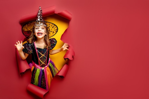 Laughing funny child girl in a witch costume of halloween looking, smiling and scares through a hole of red, yellow paper background.