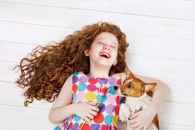 Laughing girl embracing the dog laying on a warm wooden floor.