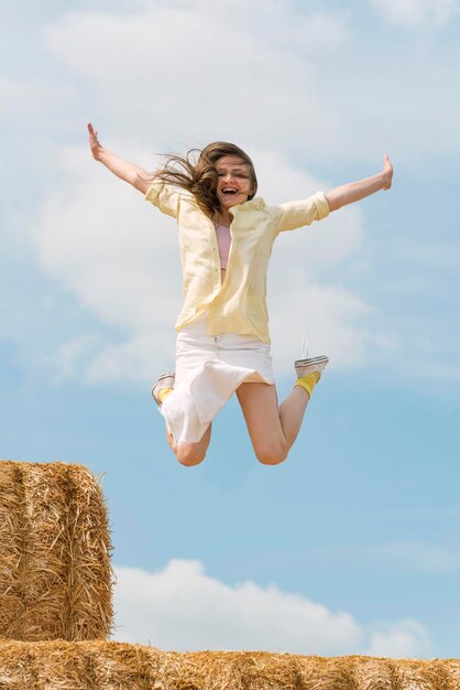 Laughing girl jumps from high haystack Happy young woman has fun on the farm on blue sky background