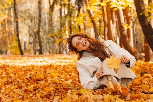 Laughing girl sits in the autumn foliage on the ground of the forest with the yellow leaves