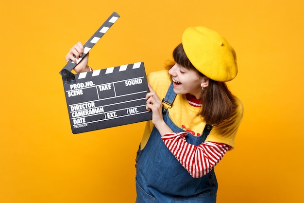 Laughing girl teenager in french beret, denim sundress looking on classic black film making clapperboard isolated on yellow background. people sincere emotions lifestyle concept. mock up copy space