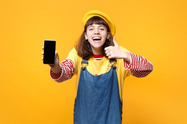 Adolescente della ragazza che ride in berretto francese che mostra il pollice in su, tenendo il telefono cellulare con lo schermo vuoto vuoto isolato sul fondo giallo della parete. persone sincere emozioni concetto di stile di vita. mock up spazio di copia.