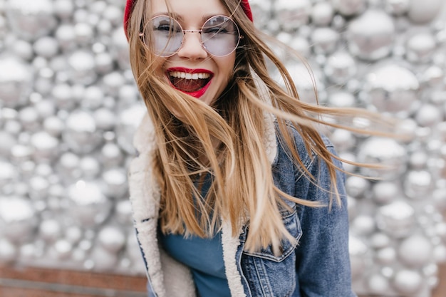 Ragazza che ride con i capelli biondi lisci in posa su sfondo scintillante incantevole modello femminile caucasico in giacca di jeans che si diverte durante il servizio fotografico