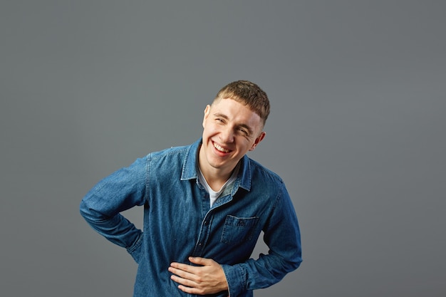청바지 셔츠를 입은 웃고 있는 남자는 회색 배경에 있는 스튜디오에서 뱃속에 손을 얹고 있습니다.