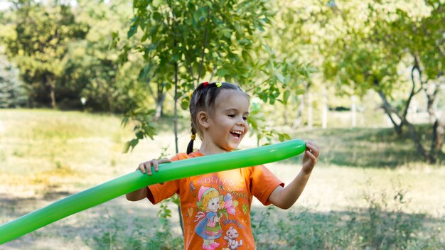 Laughing happy little girl playing with a long thin green party balloon outdoors in the park