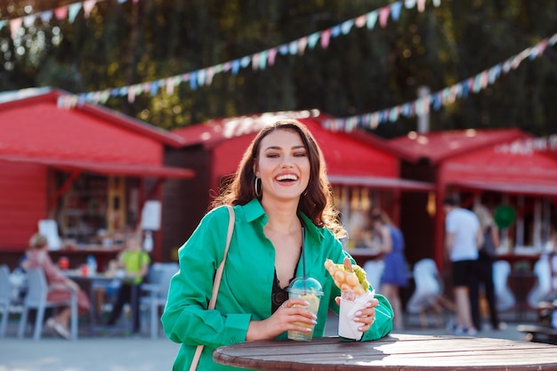 Фото Смеющаяся счастливая женщина в зеленой рубашке летом проводит время на улице на ярмарке, держа фаст-фуд