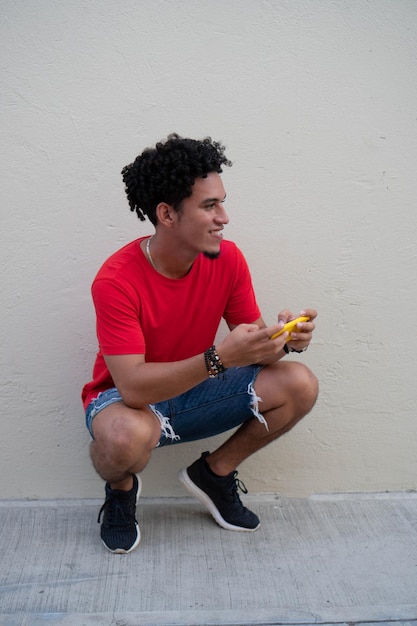 Laughing Hispanic young man crouching in front of a gray wall with a cell phone looking off into the distance