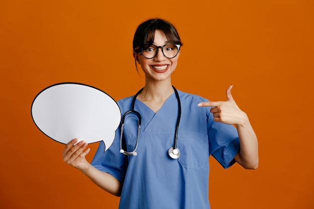 Laughing holding and points at speech bubble young female doctor wearing uniform fith stethoscope isolated on orange background