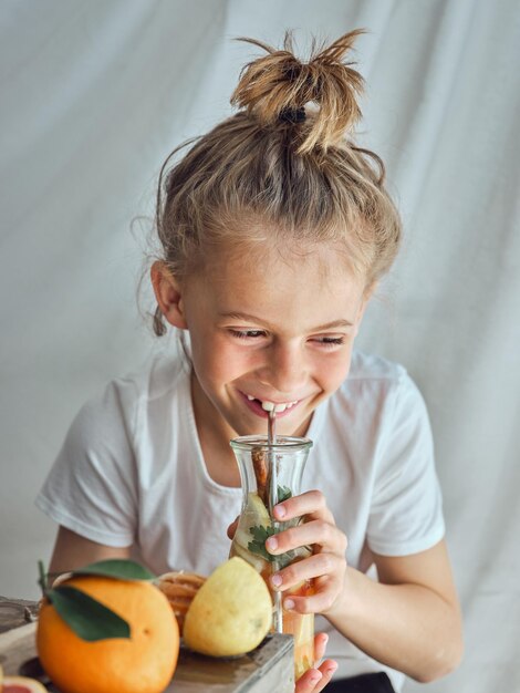 Laughing little boy in white t shirt with hair in ponytail sitting at table and drinking refreshing lemonade on white background