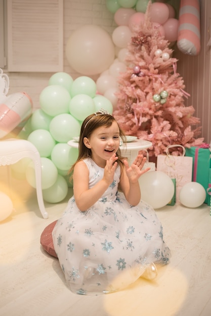 A laughing little girl in a fluffy dress sits on pillows and claps her hands in the New Year decorations