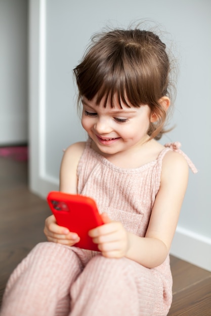 Laughing little girl with mobile phone sitting on a floor. She is playing games or watching cartoons on smartphone. Digital generation and phone addiction concept.