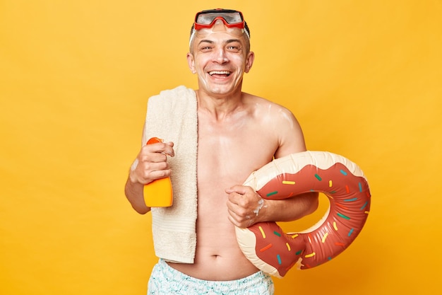 Laughing man in diving goggles and snorkel holding donut rubber ring isolated on yellow background applying sunscreen to face looking at camera