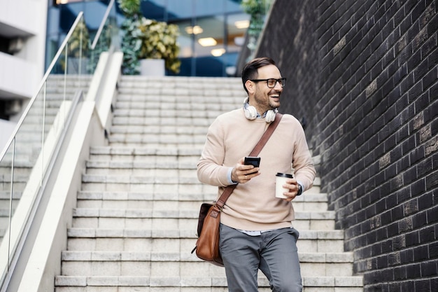 A laughing man on the stairs on street holding coffee to go and using phone