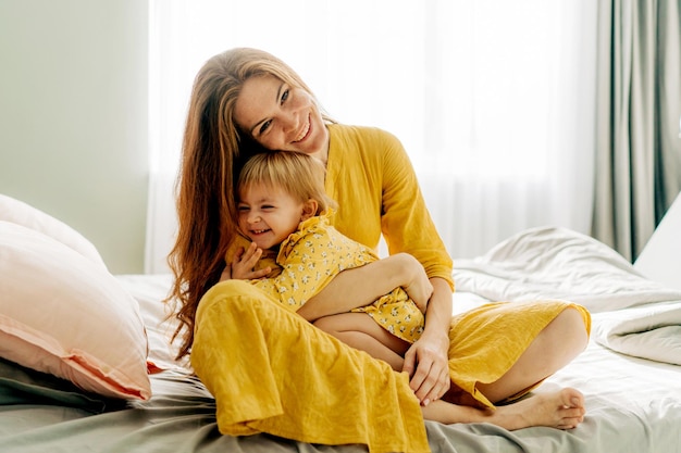 Laughing mother hugging her little daughter while sitting on the bed