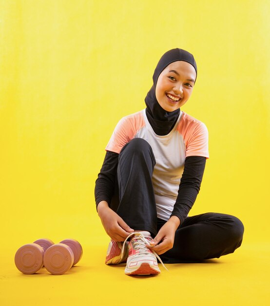 Laughing muslim woman wearing sportswear sit fixing shoelaces on yellow background person