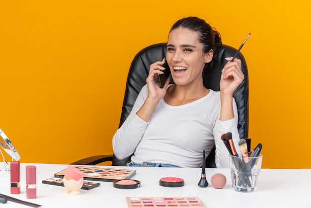 Смеющаяся красивая кавказская женщина сидит за столом с инструментами для макияжа, разговаривает по телефону и держит кисть для макияжа