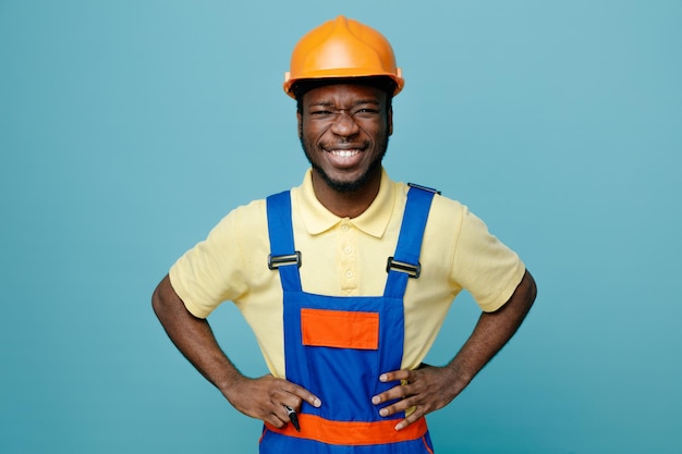 Ridendo mettendo le mani sui fianchi giovane costruttore afroamericano in uniforme isolato su sfondo blu