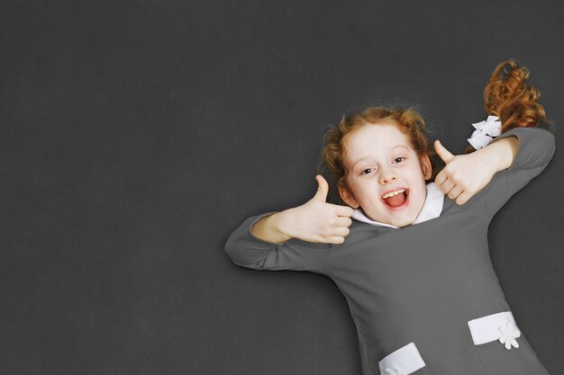 Laughing schoolgirl in grey dress showing thumbs up standing near the blackboard. 