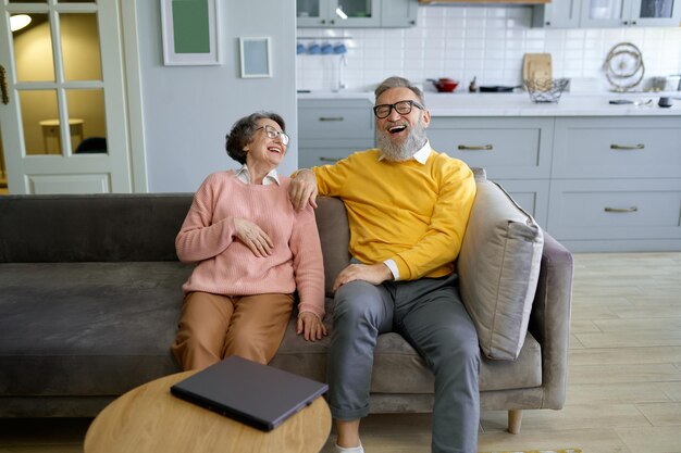 Laughing senior couple looking at each other while using laptop for shopping or booking trip online. Modern technology by elderly concept