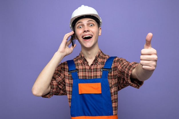 Laughing showing thumb up young male builder wearing uniform speaks on phone 