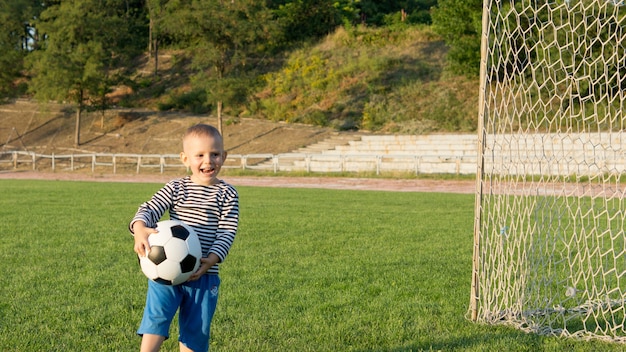 저녁 빛에 녹색 운동장에서 자신을 즐기고 그의 손에 축구 공을 가진 작은 소년을 웃고