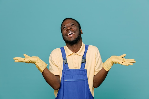 Laughing spreading hands young africanamerican cleaner male in uniform with gloves isolated on blue background