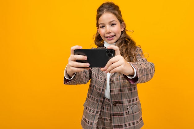 Laughing stylish blonde caucasian girl in a classic jacket takes a photo on smartphone on yellow