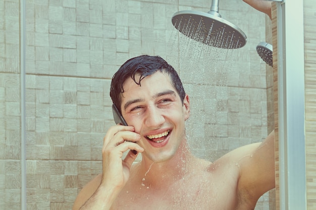 Laughing white man uses smartphone standing under streams of water pouring from shower