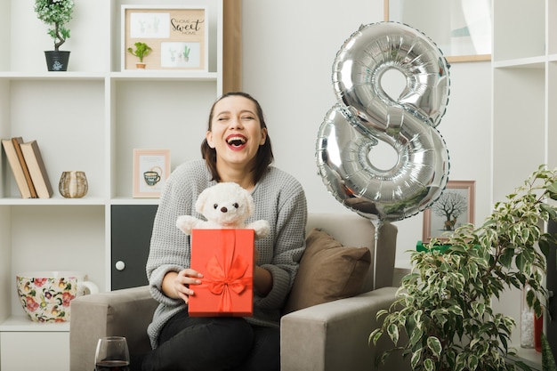 Laughing with closed eyes beautiful girl on happy women day holding present with teddy bear sitting on armchair in living room