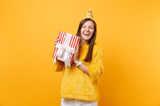 밝은 노란색 배경에서 격리된 휴가를 즐기며 축하 선물을 들고 빨간색 상자를 들고 생일 파티 모자를 쓴 웃고 있는 여성. 사람들은 진심 어린 감정, 라이프 스타일 개념입니다. 광고 영역입니다.