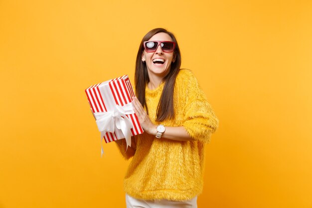 Laughing woman in red eyeglasses holding red box with gift present celebrating, enjoying holiday isolated on bright yellow background. People sincere emotions, lifestyle concept. Advertising area.