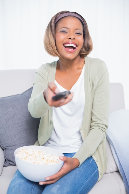 Laughing woman sitting on sofa changing tv channel while holding popcorn