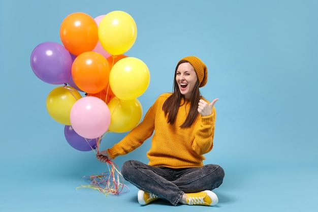 Laughing woman in sweater hat posing isolated on blue\
background. birthday holiday party, people emotions concept. mock\
up copy space. celebrating hold colorful air balloons sitting\
showing thumb up.